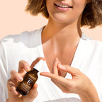 A woman in a white top holds a small amber spray bottle with a white cap near her face, showing the product in use against a soft peach background.