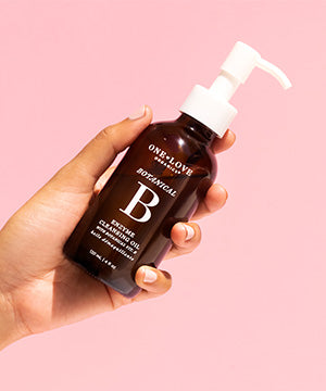 A hand holding a dark amber bottle of One Love Beauty Essentials B-Complex Cleansing Oil 