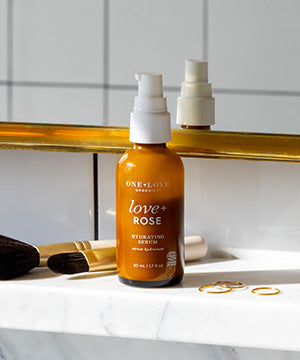 A bottle of One Love Beauty 'Love + Rose' hydrating serum stands on a white marble surface in front of a gold-framed mirror. The amber bottle has a white pump and is surrounded by cosmetic brushes and gold rings.