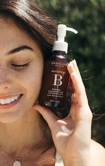 A woman holds a brown pump bottle of One Love Botanical B Enzyme Cleansing Oil 