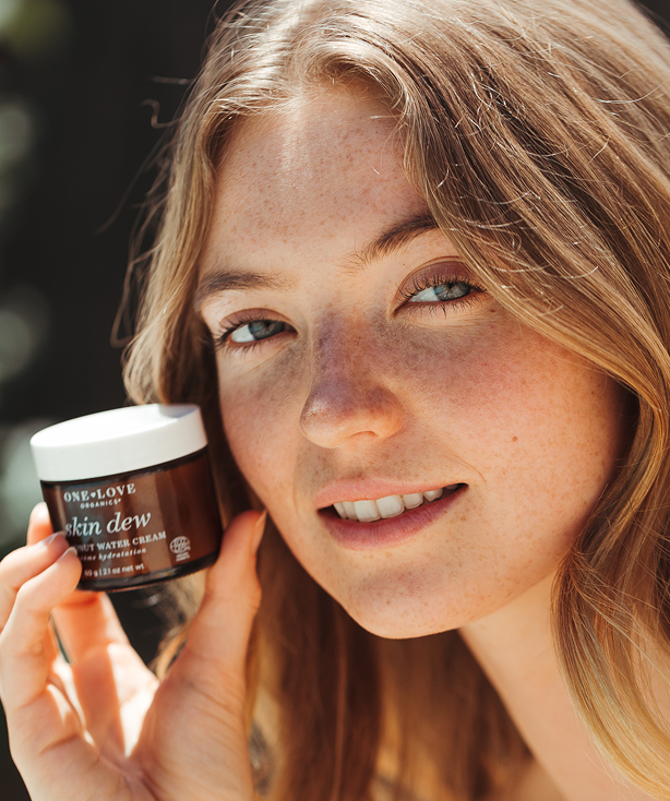 A close-up lifestyle shot of a smiling woman holding a jar of skin dew