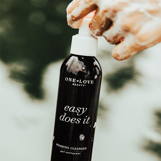A close-up of a hand applying foaming cleanser from a black pump bottle labeled 'ONE LOVE easy does it'.