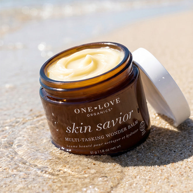 An open amber jar of One Love Organics Skin Savior Multi-Tasking Wonder Balm sits on wet sand