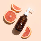 A bottle of One Love Botanicals Facial Cleanser with a white pump, styled on a light peach background with grapefruit slices.
