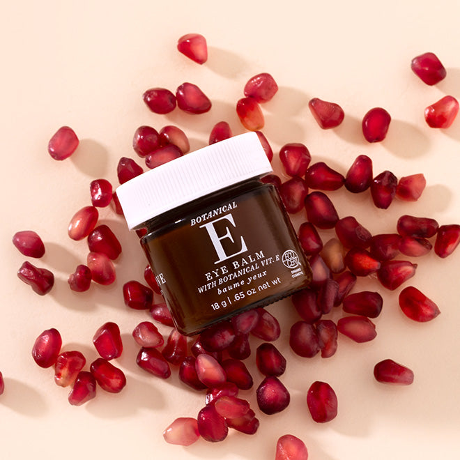 A small amber glass jar of Botanical E Eye Balm with a white lid, surrounded by scattered red pomegranate seeds on a light beige background.