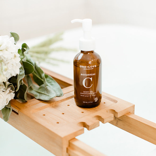 Bottle of One Love Botanical C body oil on a wooden tray with flowers in the background