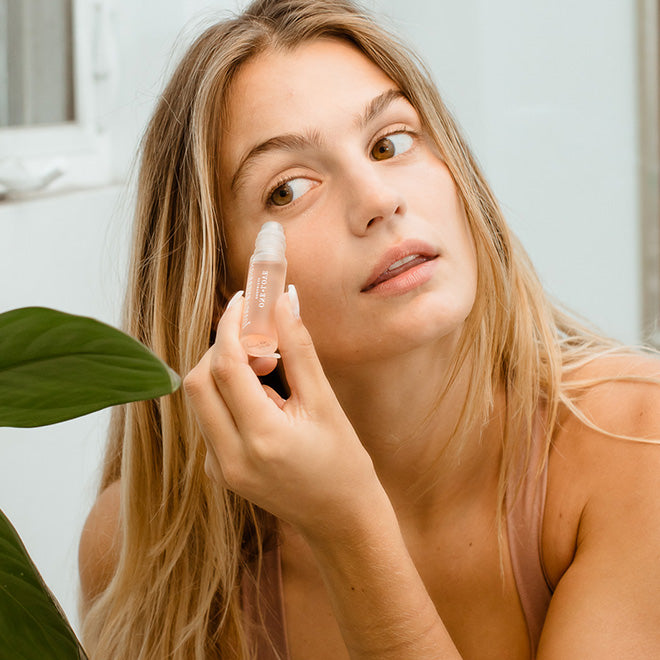 A woman with blonde hair applies a clear eyebright serum shown in a bright, clean setting with a green leaf in the foreground.