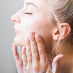 Close-up of a woman with closed eyes applying or smoothing a white facial cleanser
