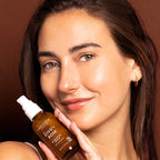 A close-up lifestyle shot of a woman holding a brown bottle of One Love Organics Love+ Rose Serum against a warm brown background.
