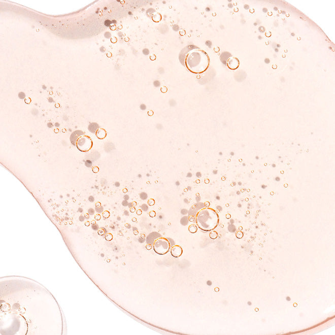 A close-up of a translucent pale pink cosmetic gel or serum with scattered gold-toned bubbles on a white background.