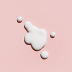 A glossy dollop of white cream or lotion on a soft pink background, with three small white spheres arranged around it.