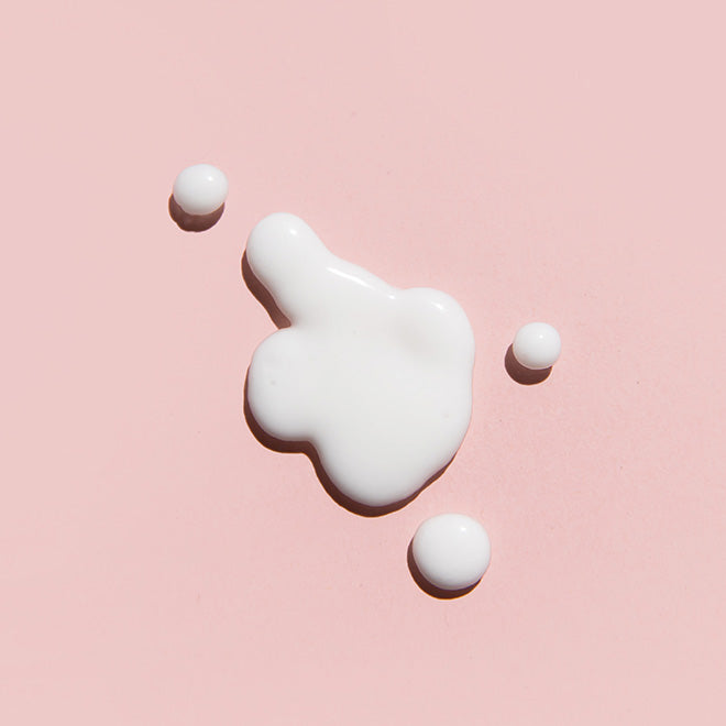 A glossy dollop of white cream or lotion on a soft pink background, with three small white spheres arranged around it.