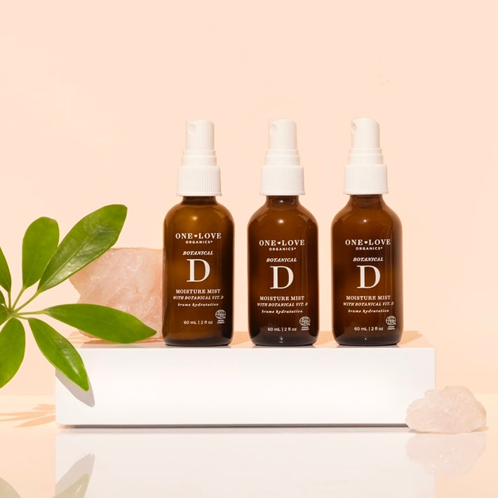 Three bottles of One Love Organics Botanical D Moisture Mist arranged on a white surface with a plant and a pink crystal in the background.
